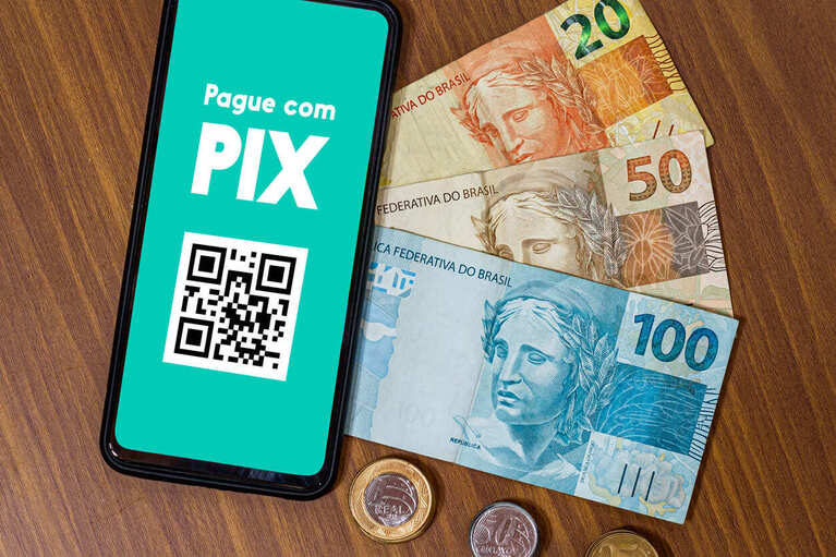 Imagem de um celular na mesa com um QR Code na tela e com a mensagem Pague com Pix. Ao lado, há notas de 20, 50 e 100 reais mais algumas moedas.