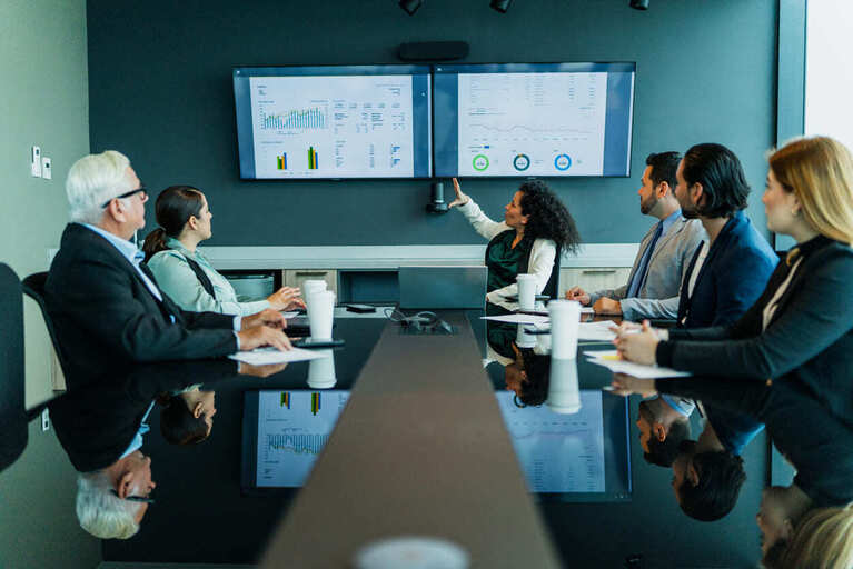Reunião de equipe em sala de conferências em uma reunião comandada por uma mulher apresentando dados em telas digitais, clima de colaboração e análise de projeções financeiras.