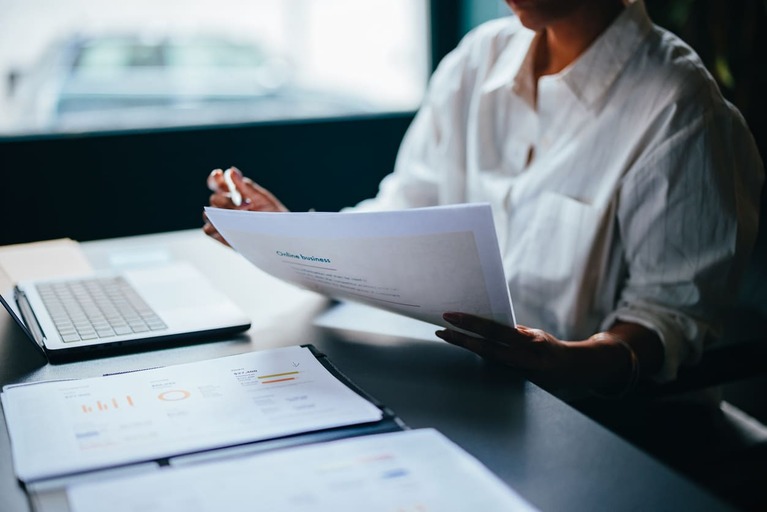 Pessoa analisando relatórios financeiros em ambiente de trabalho com computador e documentos, foco em negócios e planejamento estratégico.