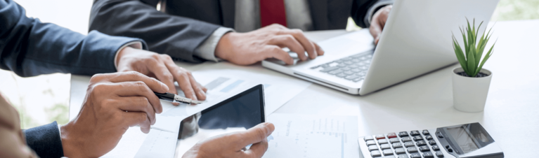  Pessoas em uma mesa com papéis, notebook, tablet e calculadora.